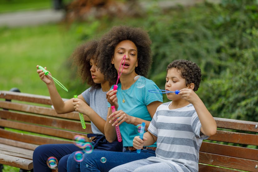 Happy African-American Family is Sitting on a Park Bench and Having a Lot of Fun While Blowing Bubbl...