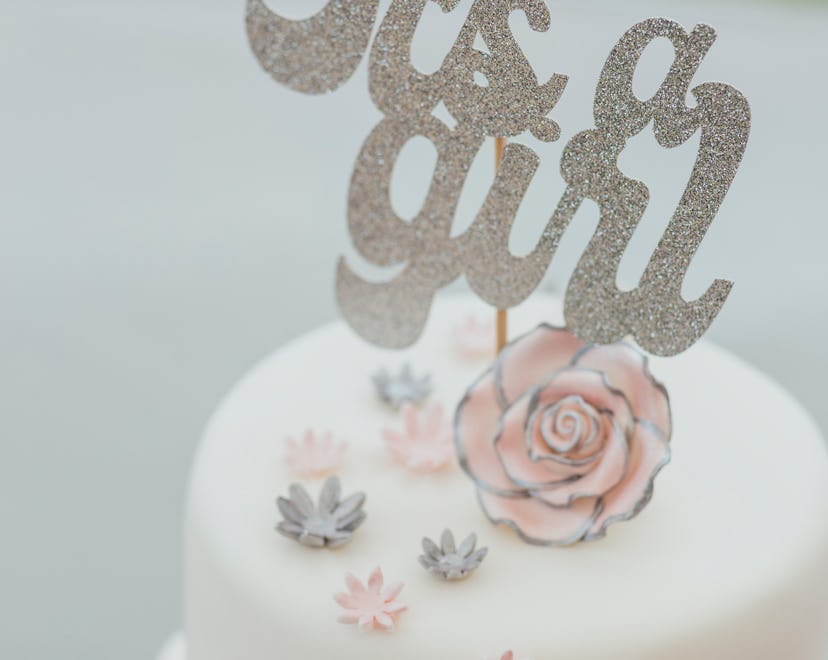 A shot of a two tier cake decorated with flowers and topped with a 'Its a Girl' sign.