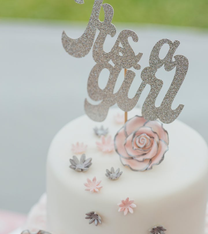 A shot of a two tier cake decorated with flowers and topped with a 'Its a Girl' sign.