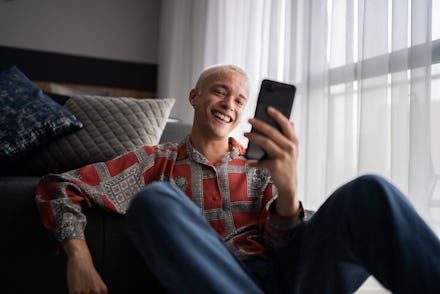 Young man using mobile phone (doing a video call or surfing on the net) sitting on the ground at hom...