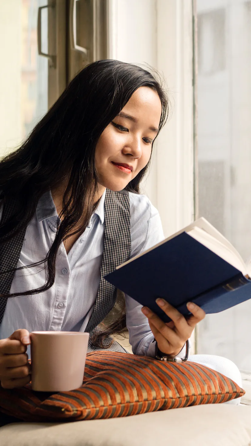 Young asian woman sitting by the window on the sofa with a cup of tea, book and pillow, laughing. Re...