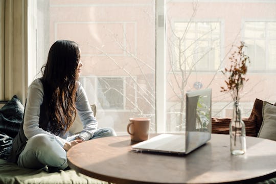 Young asian woman sitting by the window on the sofa with a laptop on a pillow in lotus position and ...