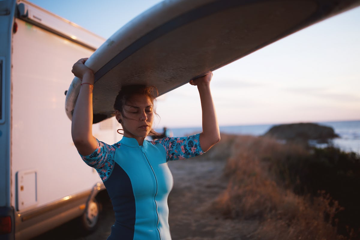 Young beautiful woman with eyes closed holding surfboard, showing the Olympic sport she'd compete in, per her zodiac sign.