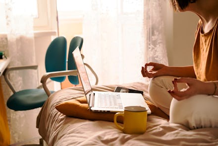 A middle-aged woman in white jeans and a yellow sweater sitting on the bed in a yoga pose in front o...