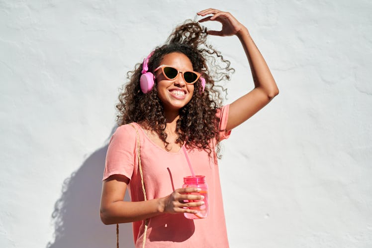 Young woman with headphones and lemonade in front of white wall in sunlight, having the best week of...