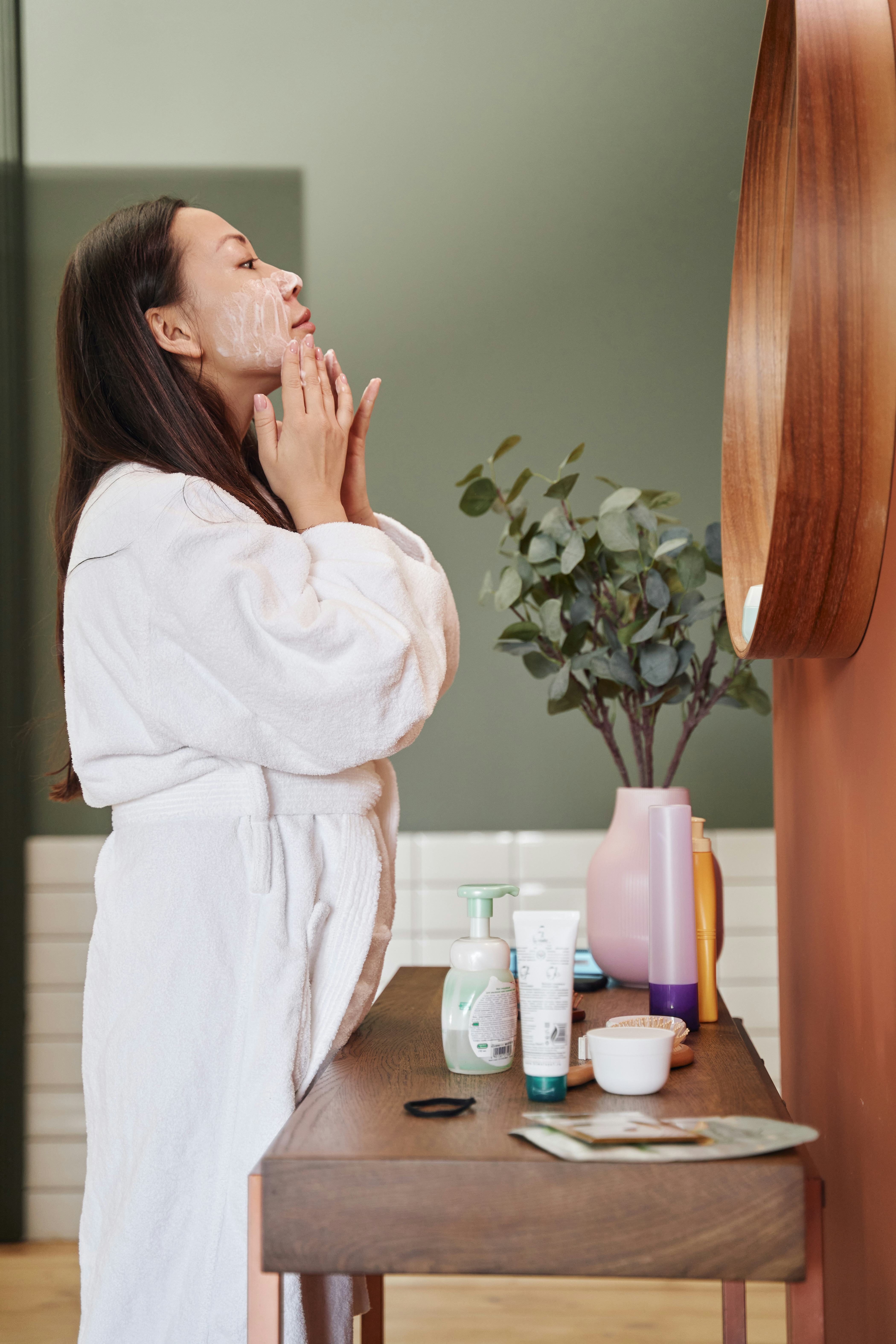 A woman standing in front of her bathroom mirror applying a face moisturizer