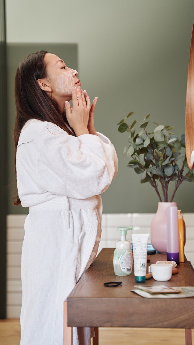 A woman standing in front of her bathroom mirror applying a face moisturizer