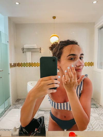 Photo of a young woman applying moisturizer and sunscreen and taking mirror selfies in the bathroom;...