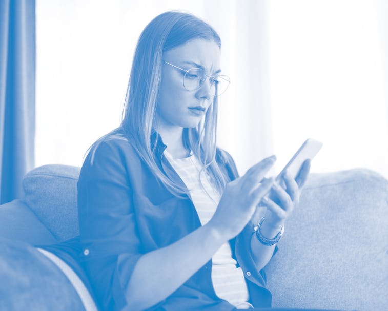 Young worried woman using social media on her phone while resting on sofa i the living room.