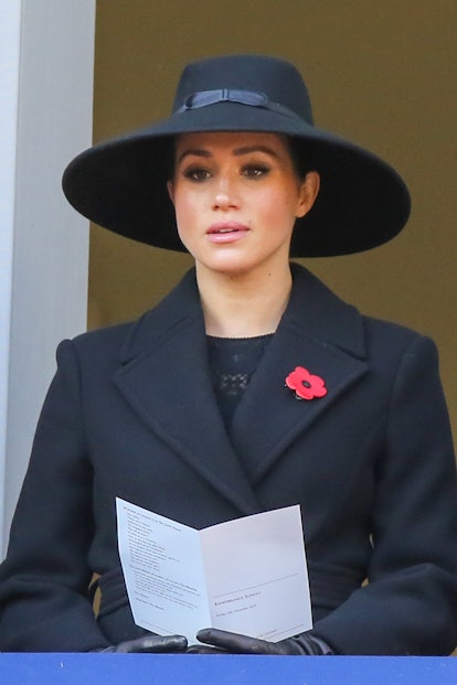 Here, Markle is shown sitting while wearing an all black ensemble consisting of a large hat, coat, a...