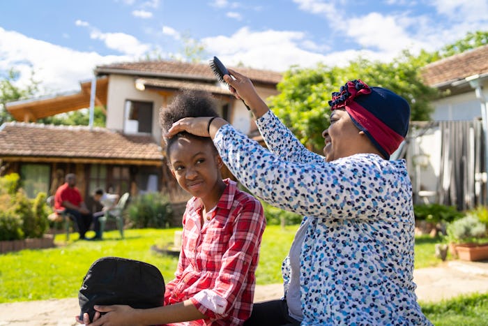 Afro-American mom is brushing her daughters' hair while they are sitting outside in the sunshine of ...