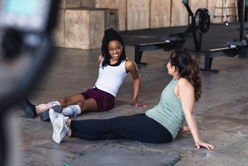 The mid adult female trainer sits on the gym floor with her young adult female client to relax after...