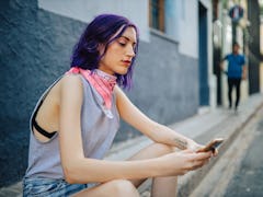 A young woman sits on the side of the street looking at her phone.