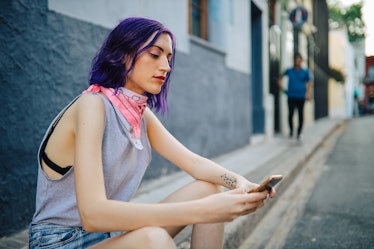 A young woman sits on the side of the street looking at her phone.