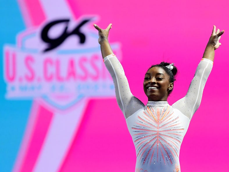 Simone Biles is shown wearing a embellished white leotard while standing in front of a pink and blue...