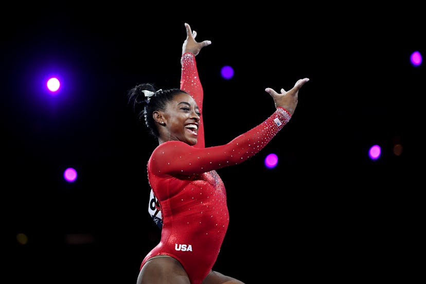 Simone Biles is wearing a red leotard and is smiling while performing at a competition.