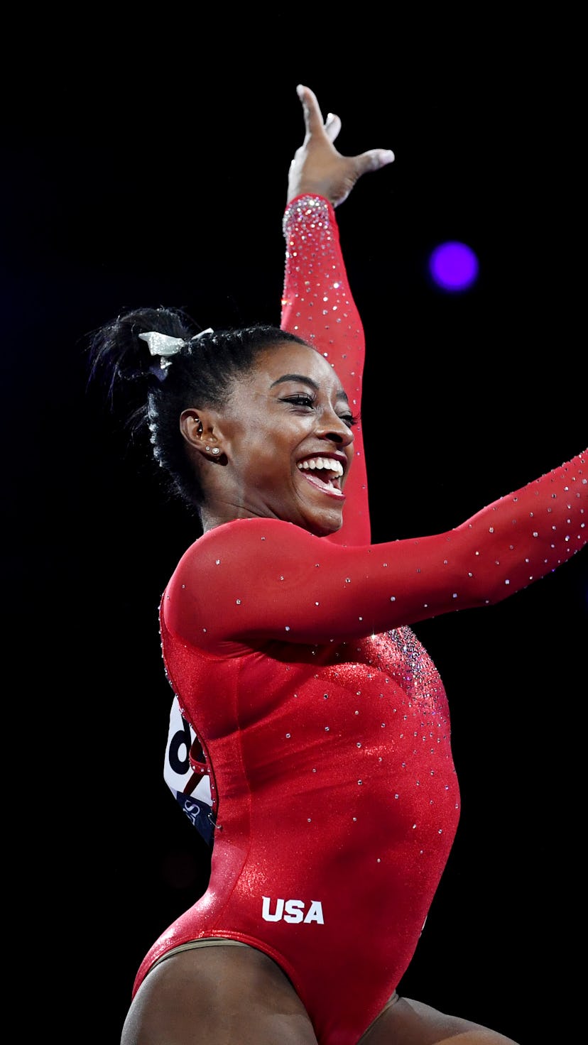 Simone Biles is wearing a red leotard and is smiling while performing at a competition.