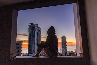 A woman sitting on her balcony ledge alone looking out at the city view at sunrise