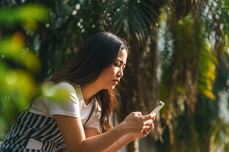 A young woman is using a smart phone among palm tree leaves.