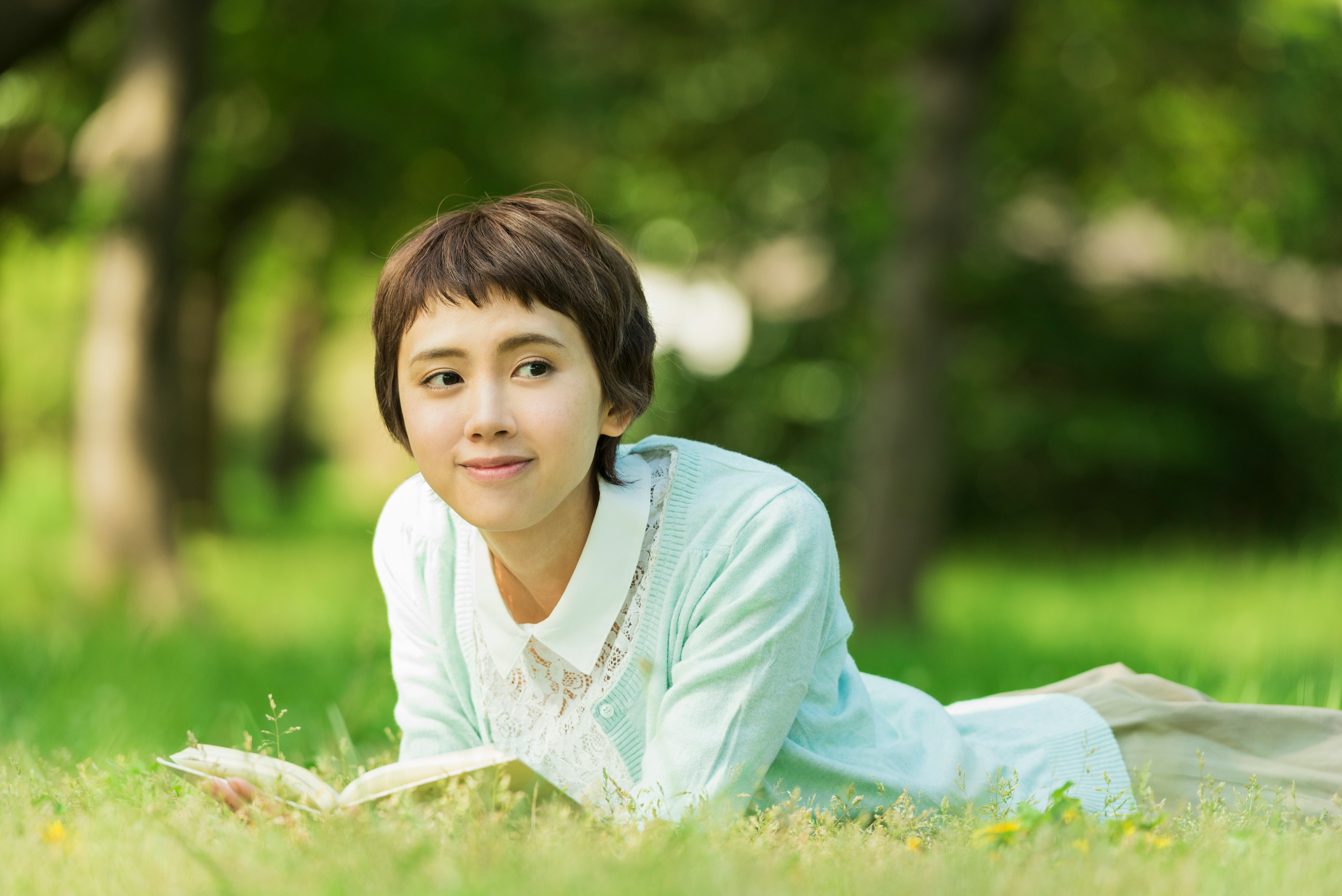 A smiling, femme Asian person with short hair lies in the grass reading a book.