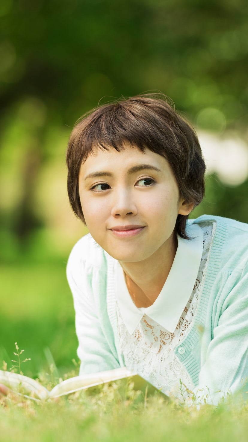 A smiling, femme Asian person with short hair lies in the grass reading a book.