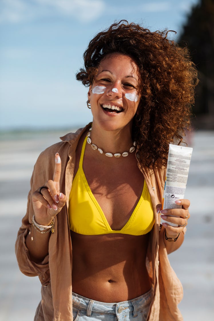 A tan young woman with curly hair laughs while applying sunscreen to her face on the beach
