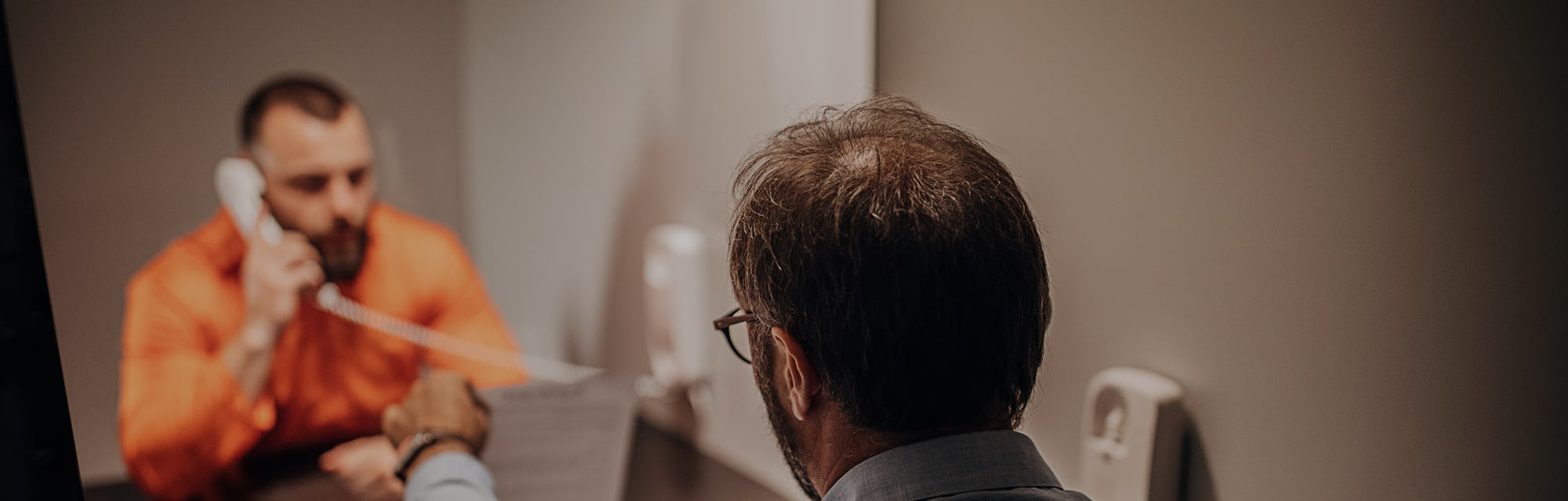 Two men, male prisoner talking over the phone with his lawyer in prison visit room.