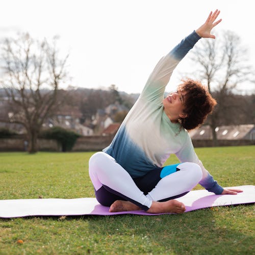 A person wears a sweater while doing yoga on a mat in a park. Working out while you're sore can help...