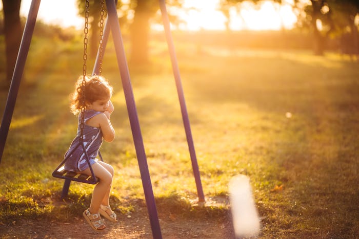 Cute little lonely sad girl swinging at sunset in public park.