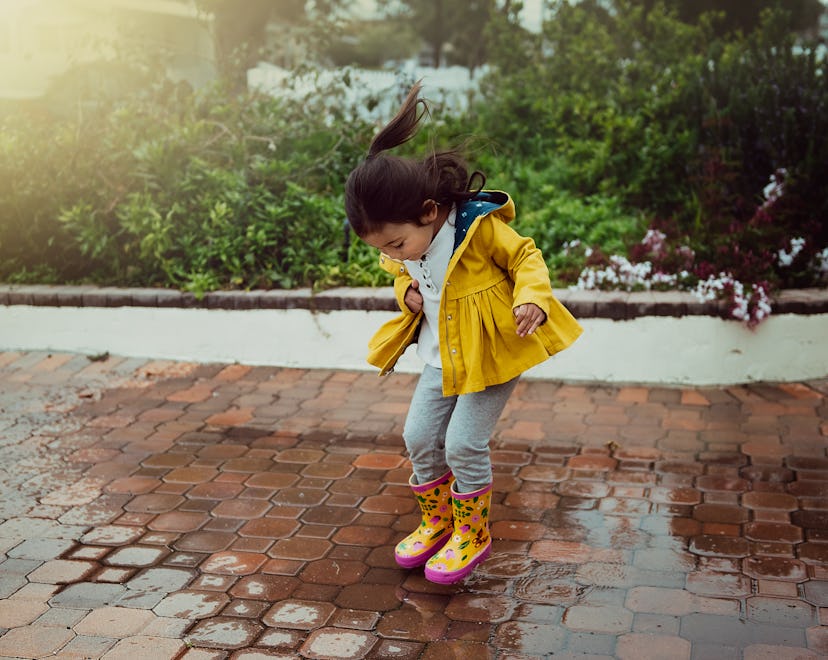 A three year old girl in a yellow jacket and flowered yellow and pink boots jumps in a puddle.