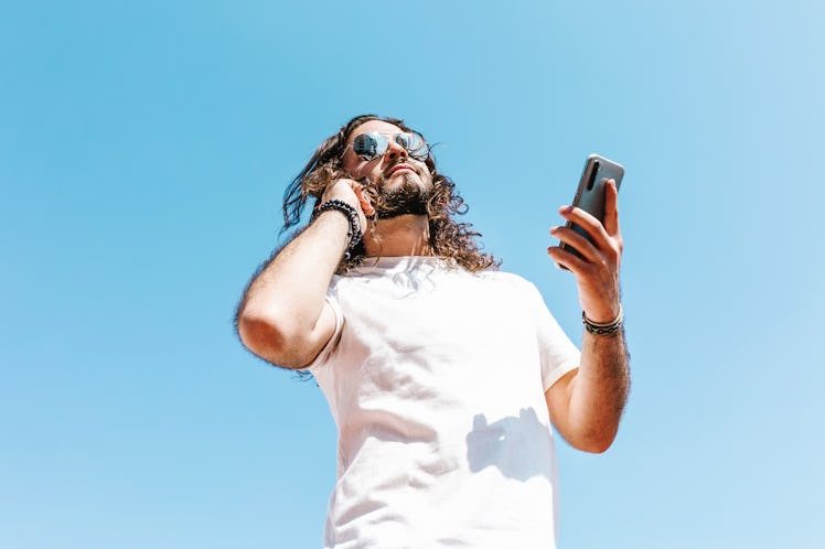 From below of bearded Latin American male in white t shirt and modern sunglasses with cellphone on b...