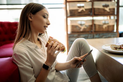 A person sits on the floor in front of their couch, texting and eating a sandwich. Reaching out to r...