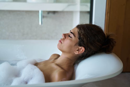 Side view attractive relaxed female with eyes closed enjoying hot foamy water in bathtub in contempo...