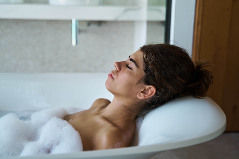 Side view attractive relaxed female with eyes closed enjoying hot foamy water in bathtub in contempo...
