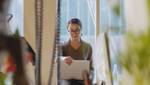 Stock image of a young Chinese Asian woman sitting by an office window as she opens her laptop.
