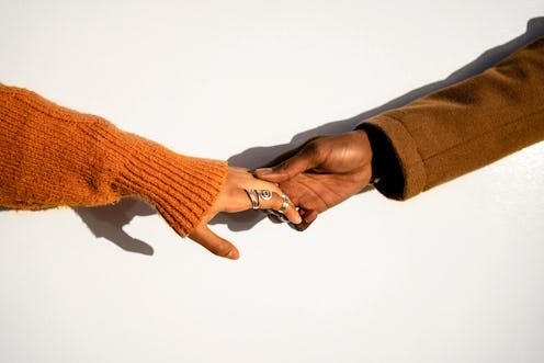 Crop woman in orange sweater and rings holding hand of black boyfriend in brown coat against wall in...