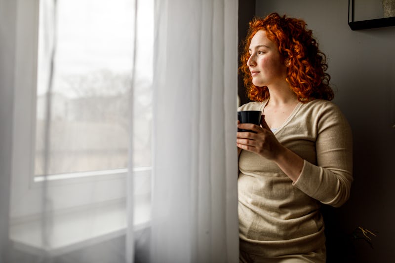 Copy space shot of smiling young woman standing by the window in her living room, looking outside an...