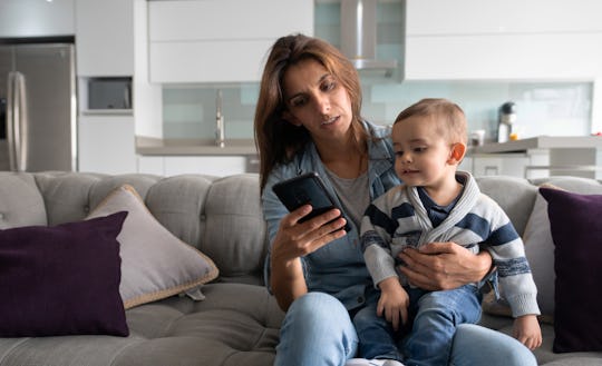 Working Latin American mother staying at home and holding her baby while texting on her cell phone -...