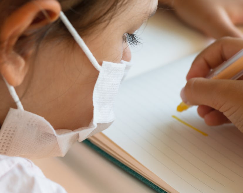 Over the shoulder  view of child with face mask is writing with yellow pen.