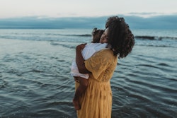 Woman standing and holding her daughter lovingly in front of the ocean with an overcast sky.