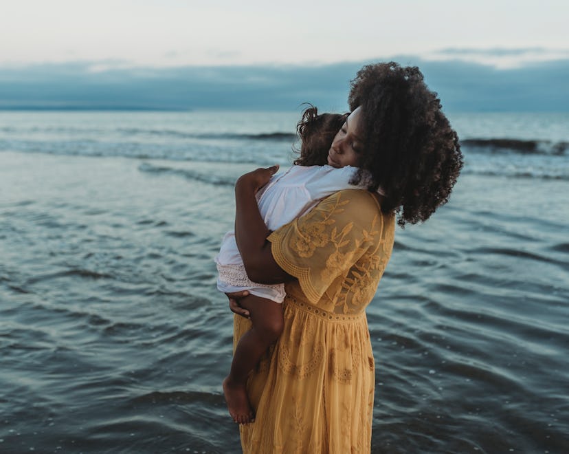 Woman standing and holding her daughter lovingly in front of the ocean with an overcast sky.