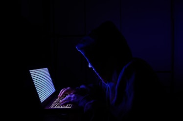 A person dressed as an internet hacker in front of a laptop with a binary code displayed on screen 