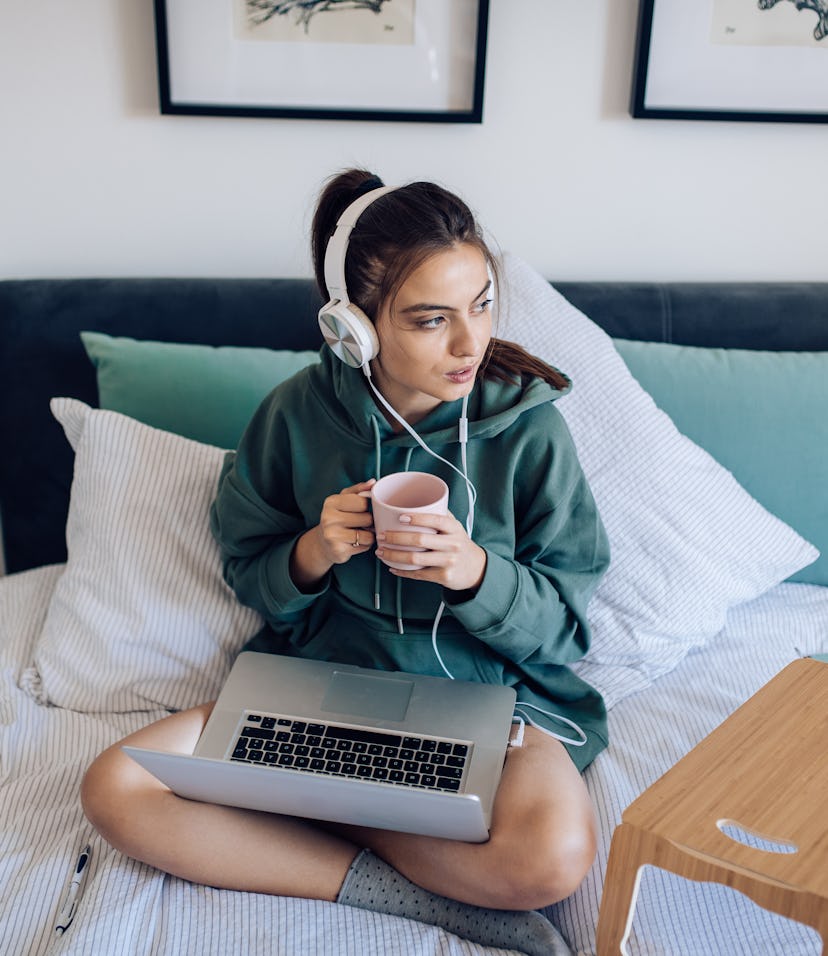 A college student drinks coffee and sits with their laptop, in their dorm room that feels like a hot...