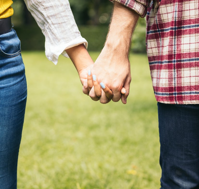 Anonymous man and woman holding hands while standing on grassy lawn on summer date in park