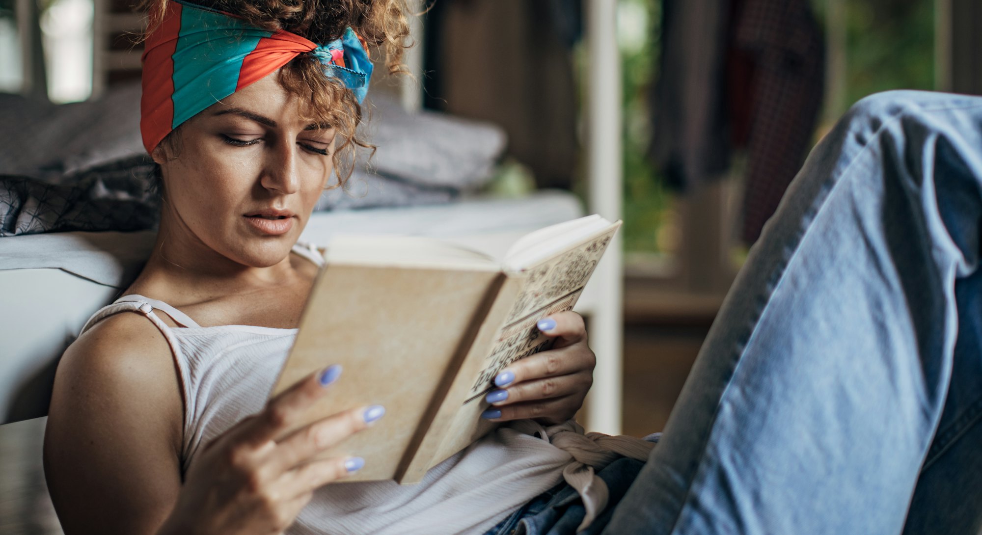 One woman sitting on the floor in bedroom at home. She is reading a book.