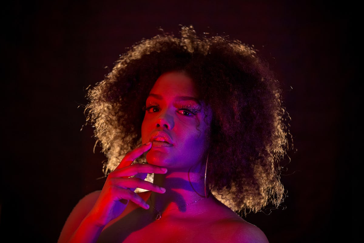 Jovem mulher tocando lábios e olhando para a câmera sob luz brilhante contra fundo preto durante t ...