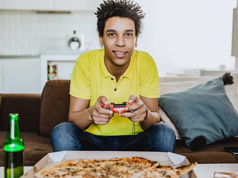 Young African American man eating pizza, drinking beer and playing video games