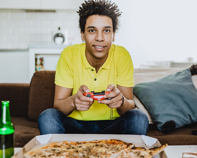 Young African American man eating pizza, drinking beer and playing video games