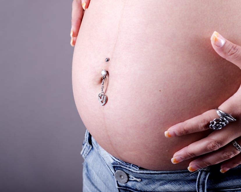 Horizontal studio shot on gray of pregnant woman's belly with piercing and hands.