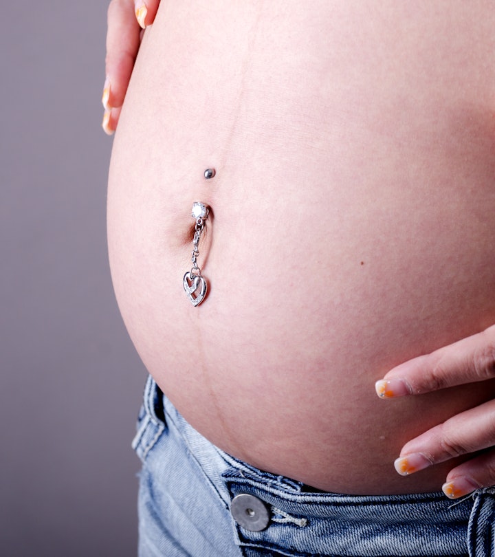 Horizontal studio shot on gray of pregnant woman's belly with piercing and hands.